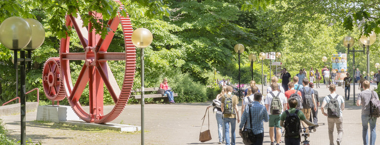 Kopfbild Studenten rote Zahnräder Campus Nord Studenten im Sommer auf dem Campus Nord, rote Zahnräder im Hintergrund